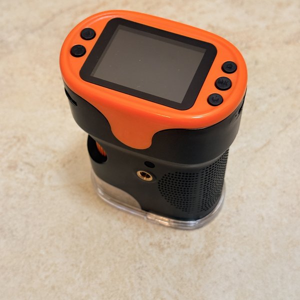 1000X digital microscope with a bright orange plastic housing around a small 2-inch black screen and side control buttons, resting on a cream limestone counter